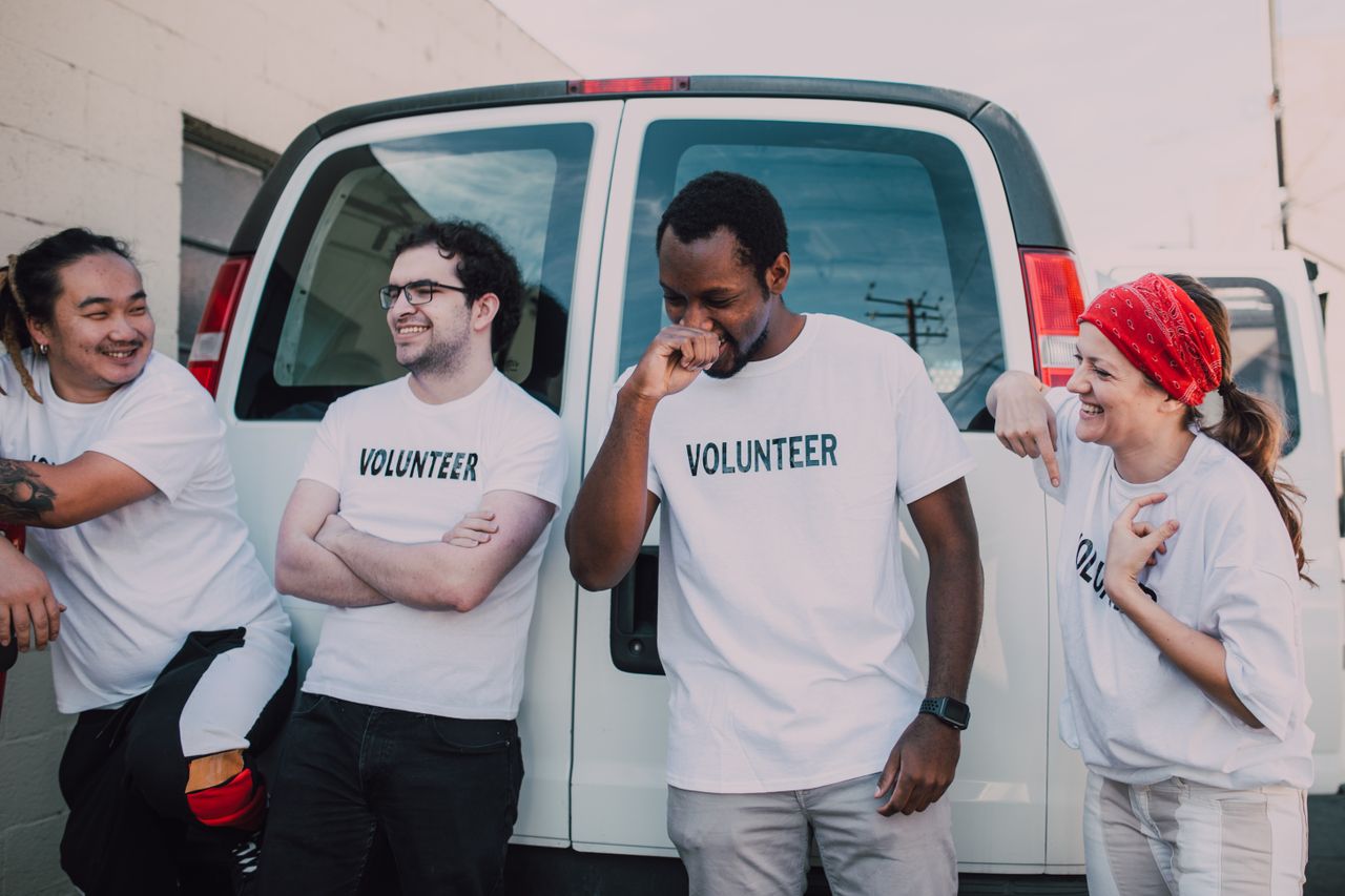 Group of cheerful volunteers wearing white shirts, leaning against a van outdoors.
